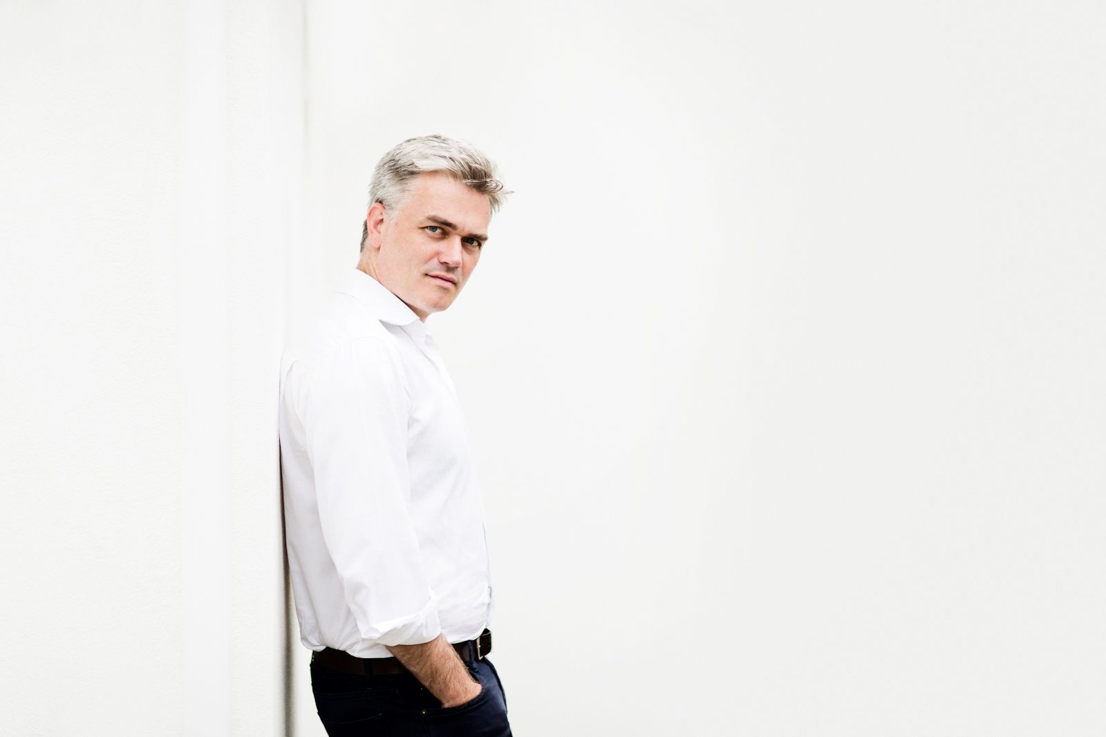 Photo of conductor Ed Gardner in a white shirt leaning against a white wall