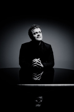 Black and white image of Benjamin Grosvenor leaning on a piano