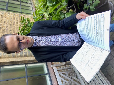 Image shows a young man with dark hair and glasses reading a large music score