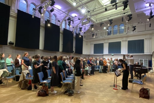 Image is a photograph of a choir rehearsing in a large hall