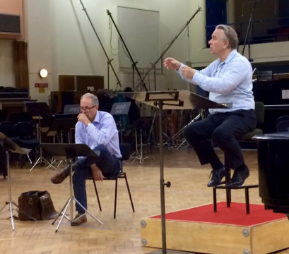 Image is of two men in blue shirts, both sat behind music stands conducting a choir